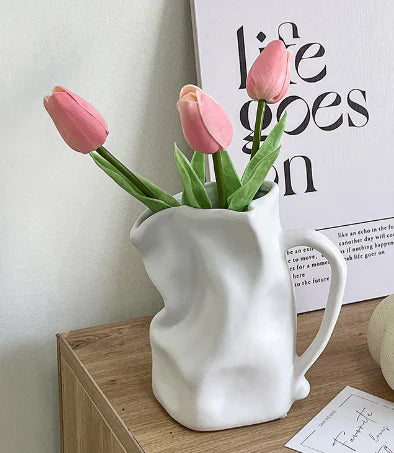 White ceramic pitcher with pink tulips on a wooden surface, with a 'Life Goes On' poster in the background.