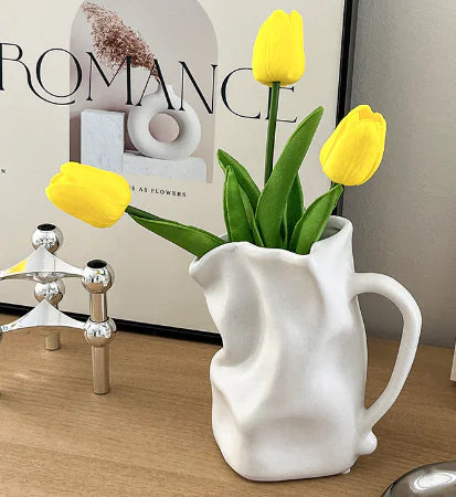 White pitcher-shaped vase with yellow tulips on a wooden surface.