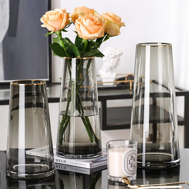 Three glass vases with a candle and flowers on a reflective surface.