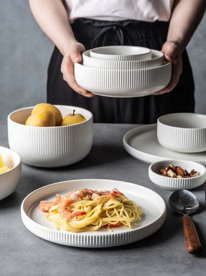 Person holding a stack of white ceramic bowls with a table setting featuring pasta and fruit.