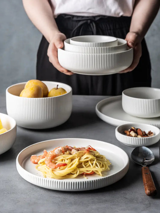 Person holding a stack of white ceramic bowls with a table setting featuring pasta and fruit.