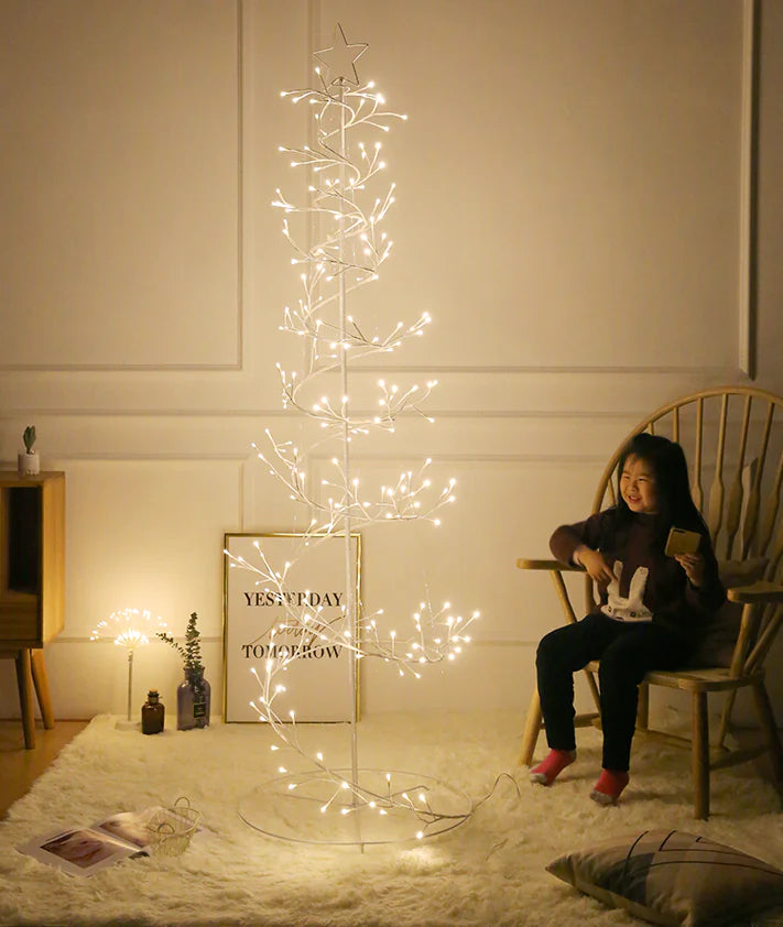 Woman sitting next to a decorative light tree in a room with a white wall and wooden furniture.